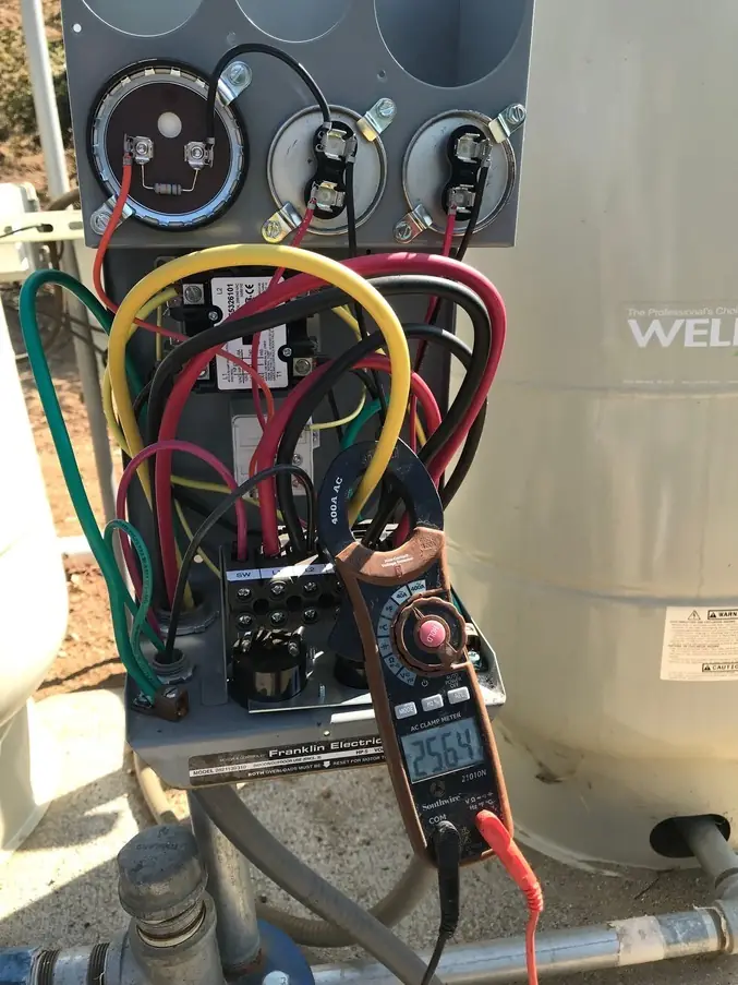 A clamp meter reading amperage on the wires of a well pump control box next to a pressure tank