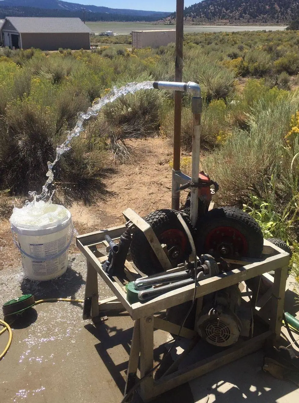 Water discharging from a test pipe during a well flow test, beside a bucket and a hose reel