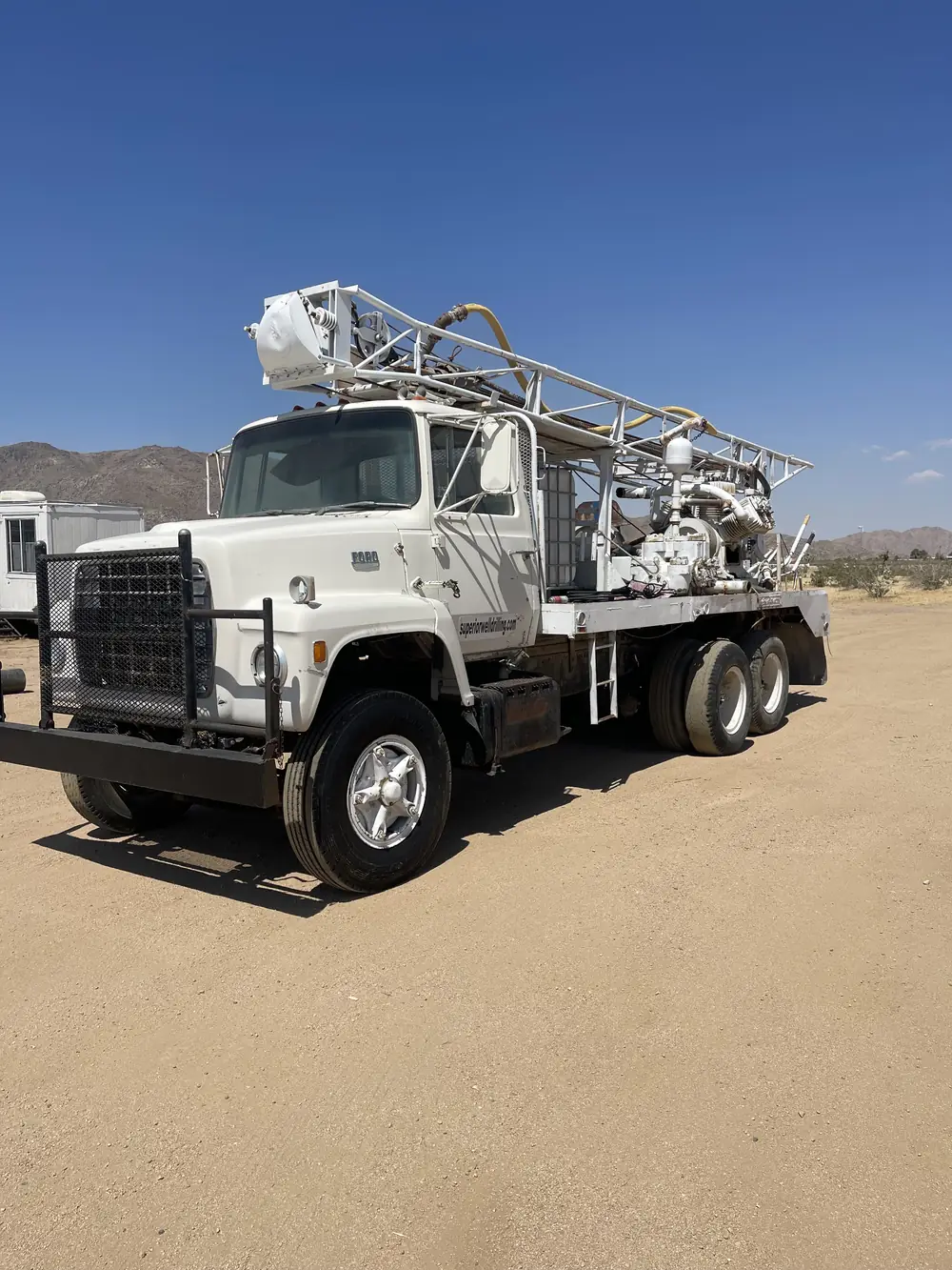 A white Ford drilling rig truck on a dirt pad in the High Desert under a clear blue sky